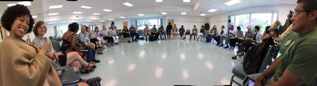 Panoramic view of a large meetign room with 40+ attendees seated closely in a circle. At the left-hand side of the image is a young (20s) light-skinned, Black actress with curly black hair, wearing a tan, oversized sweater, smiling into the camera, next to a middle-aged white woman, with a short-bob hair cut who grins. ON the other side of the circle, in the right-hand corner of the image is a middle-aged Native American man, with shortly cropped dark hair, wearign glasses and an olive green t-shirt, lookign out across the circle. Florescent lights are seen on the ceiling and reflectign off the highly polished white floor. Windows around the room allow bright natural light. 