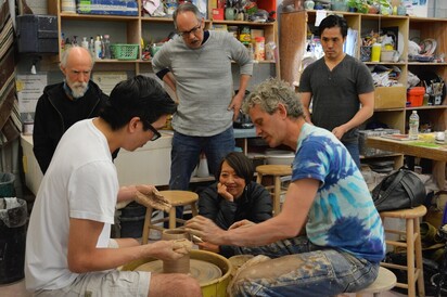 IN the bottom, center of the image master potter Bob Deane sits at a pottery wheel, wearing a blus tie-dye shirt and long pants covered in clay across from an actor in a white t-shirt with thick black-frame glasses and dark black hair. Playwrithg Jeanne Sakata, a woman kneels with her hand on her cheek behind and between the men at the pottery wheel, whe looks on with great interest and delight. Three men stand around the pottery wheel toward the back of the image studying the work on the wheel. The room is a pottery studio with shelves stocked full of materials and supplies all across the wall. 