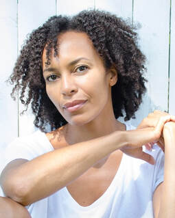 A headshot of playwright Eisa Davis, a light-skiined black woman in her 40s, with tight dark curls hanging, who gazes casually into the camera one arm across her body connecting with her other wrist. She wears a v-neck white t-shirt and is leanign up against a white wall with thich wooden slats.  