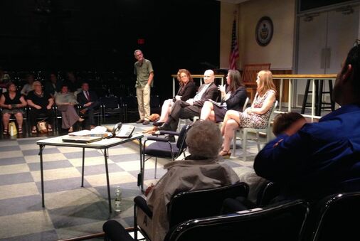 A horizontal image of a panel with four speakers dressed professionally seated on a theatre stage that is set in a court room, the facilitator a middle-aged white man stands to the side in the background, and audience members are on both sides of the panel, in the distance, focused on the conversation, seated on the far-side of the image, and in the foreground of the image to the right-hand side. 